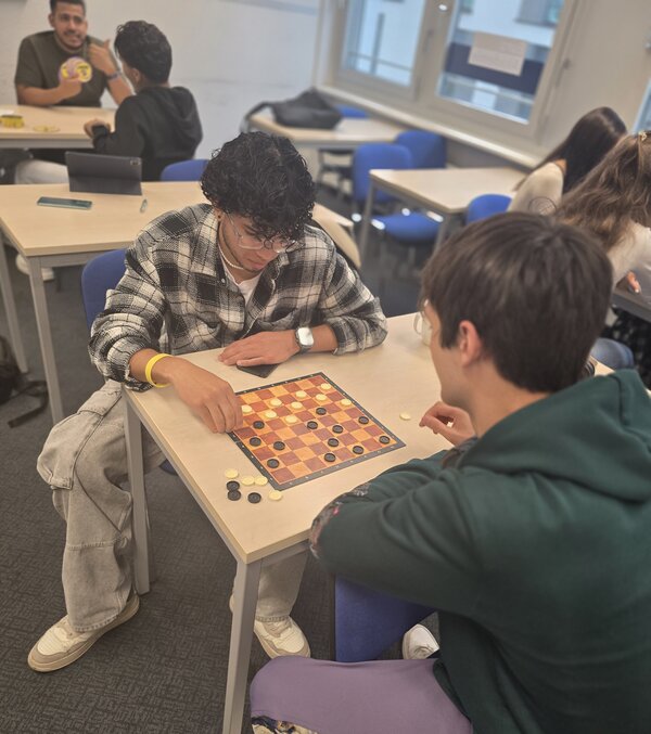 Spielen, Sprechen, Spaß haben – Kreative Gesprächsanlässe im Jugendintegrationskurs Zwei junge Männer sitzen an einem Tisch und spielen Dame. Beide konzentrieren sich auf das Spielbrett, während im Hintergrund weitere Personen sitzen und sich unterhalten.