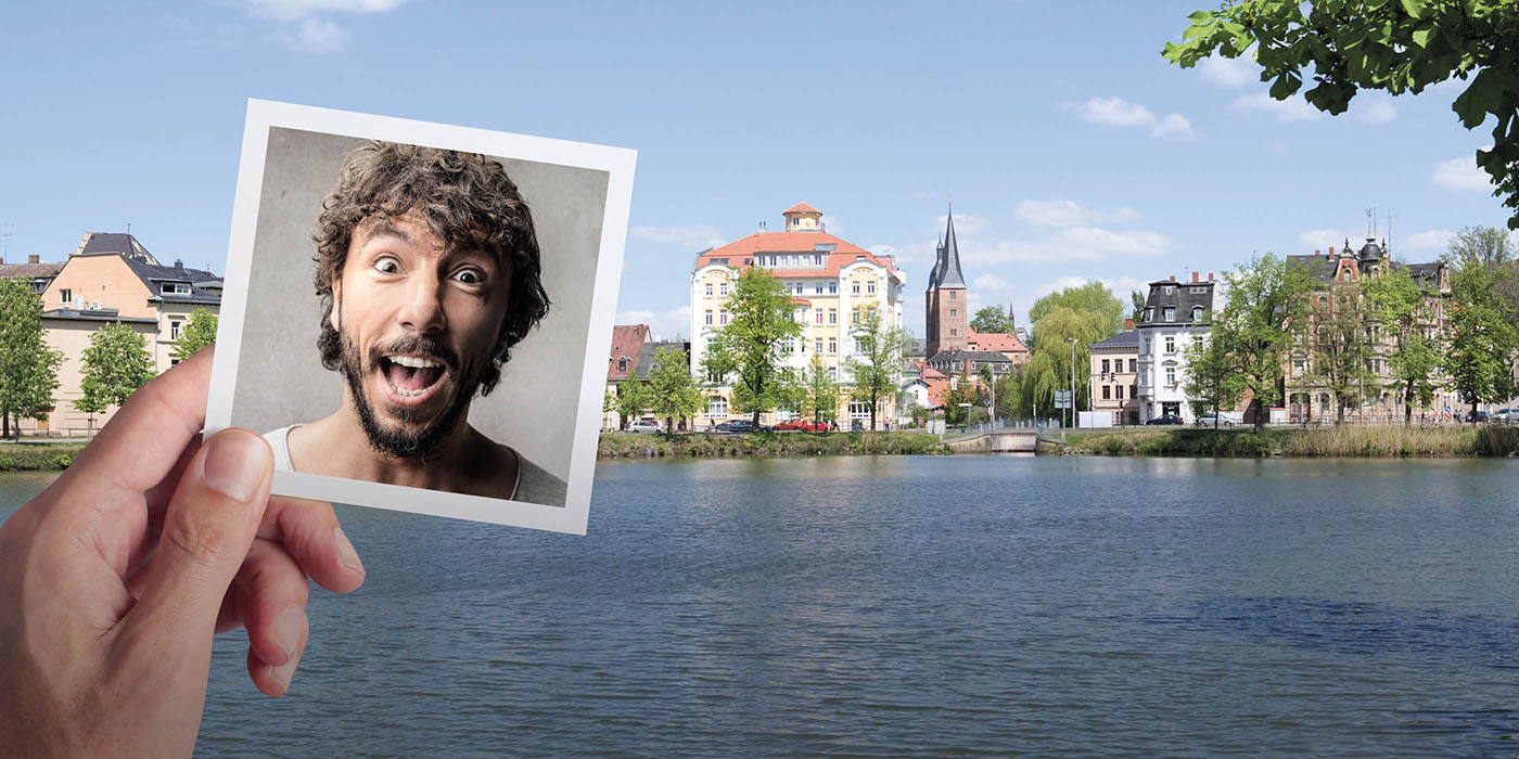 Eine Hand hält ein Foto von dem Gesicht einer überrascht schauenden Person vor eine malerische Ansicht von Gebäuden, Bäumen und einem Gewässer im Vordergrund. Es handelt sich um eine Aussicht auf die Stadt Altenburg.