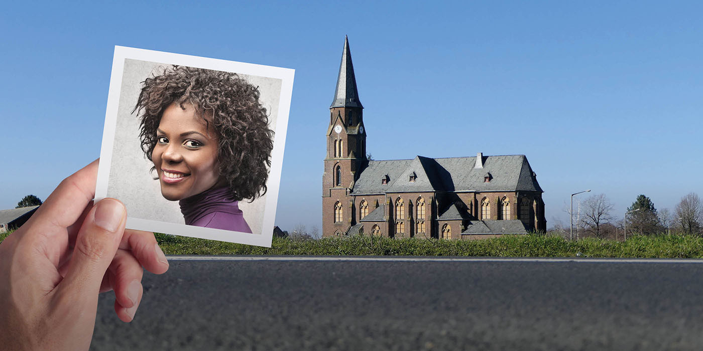 Eine Hand hält ein Foto von dem Gesicht einer lächelnden Person vor eine Ansicht einer Kirche. Im Vordergrund ist eine asphaltierte Straße zu sehen.