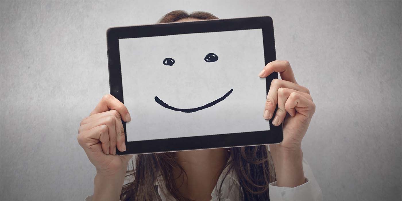 Eine Person hält einen Monitor vor das Gesicht, auf dem ein Smiley zu sehen ist, dargestellt mit zwei Punkten und einem gebogenen Strich.